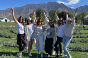 Seis mujeres sonriendo y posando con las manos por encima de la cabeza, rodeadas de vegetación.