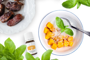 Eine Schale mit frischer Mango, Joghurt und Kokosnussraspeln neben Datteln, frischem Basilikum und ätherischem Plusöl Lemon+