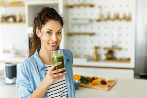 Frau in heller, ordentlicher Küche mit einem grünen Smoothie im Glas in der Hand und frischem Obst im Hintergrund