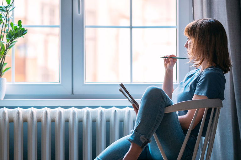 Bild einer Frau, die am Fenster sitzend in ihr Tagebuch schreibt.