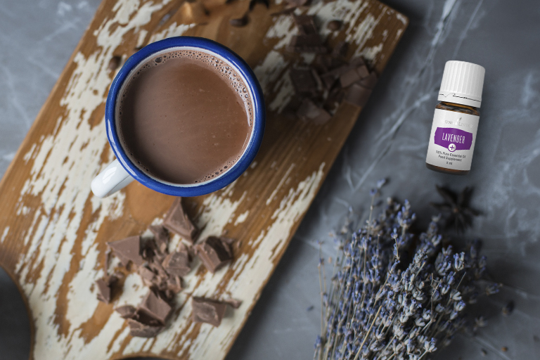 Imagen de un chocolate caliente rodeado de trozos de chocolate, ramitos de lavanda y un frasco de aceite esencial Lavender+.