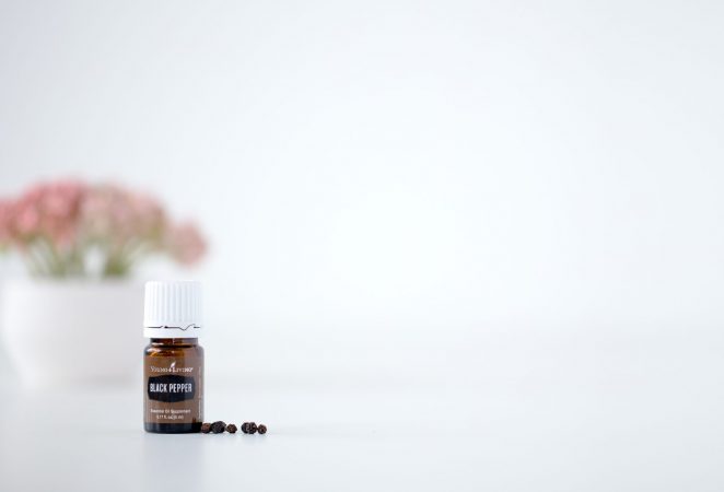 a bottle of black pepper essential oil on a white background with a few peppercorns and a bouquet faded in the background